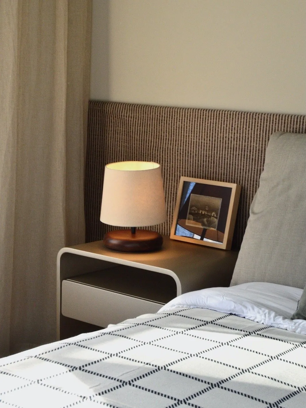 Mesita de luz con una lámpara, un libro y un cuadro, junto al respaldo de la cama